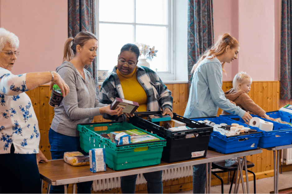 A community fridge and pantry event. The photo is taken in a community hall. 5 people from children to an old woman are looking at the surplus food and surplus products and are selecting items to take home. All items are dry goods stored in baskets on the table. 