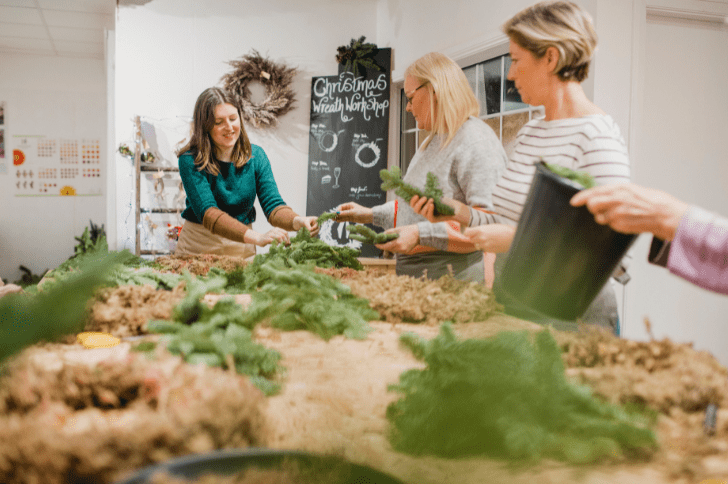 In the centre is a group of women around a worktop making sustainable Christmas wreaths out of natural products and foliage.